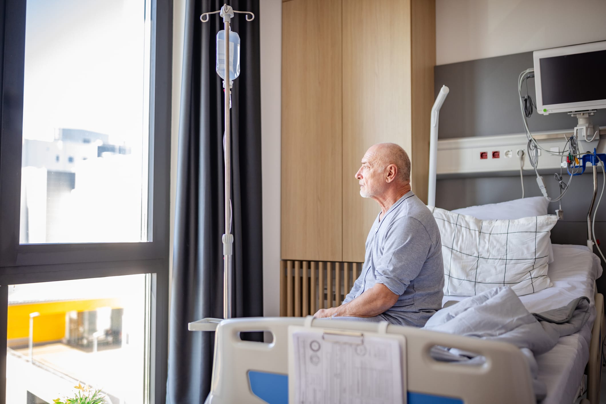 In a hospital setting, an elderly man sits up in his bed, looking out the window.